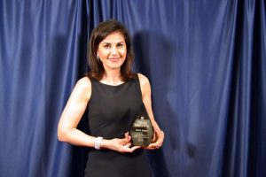 A woman in a black dress holds an award statuette in front of a blue curtain