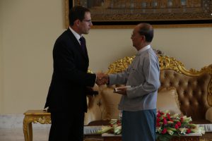 Two men shaking hands, one in a western style suit, the other in traditional Burmese clothes.